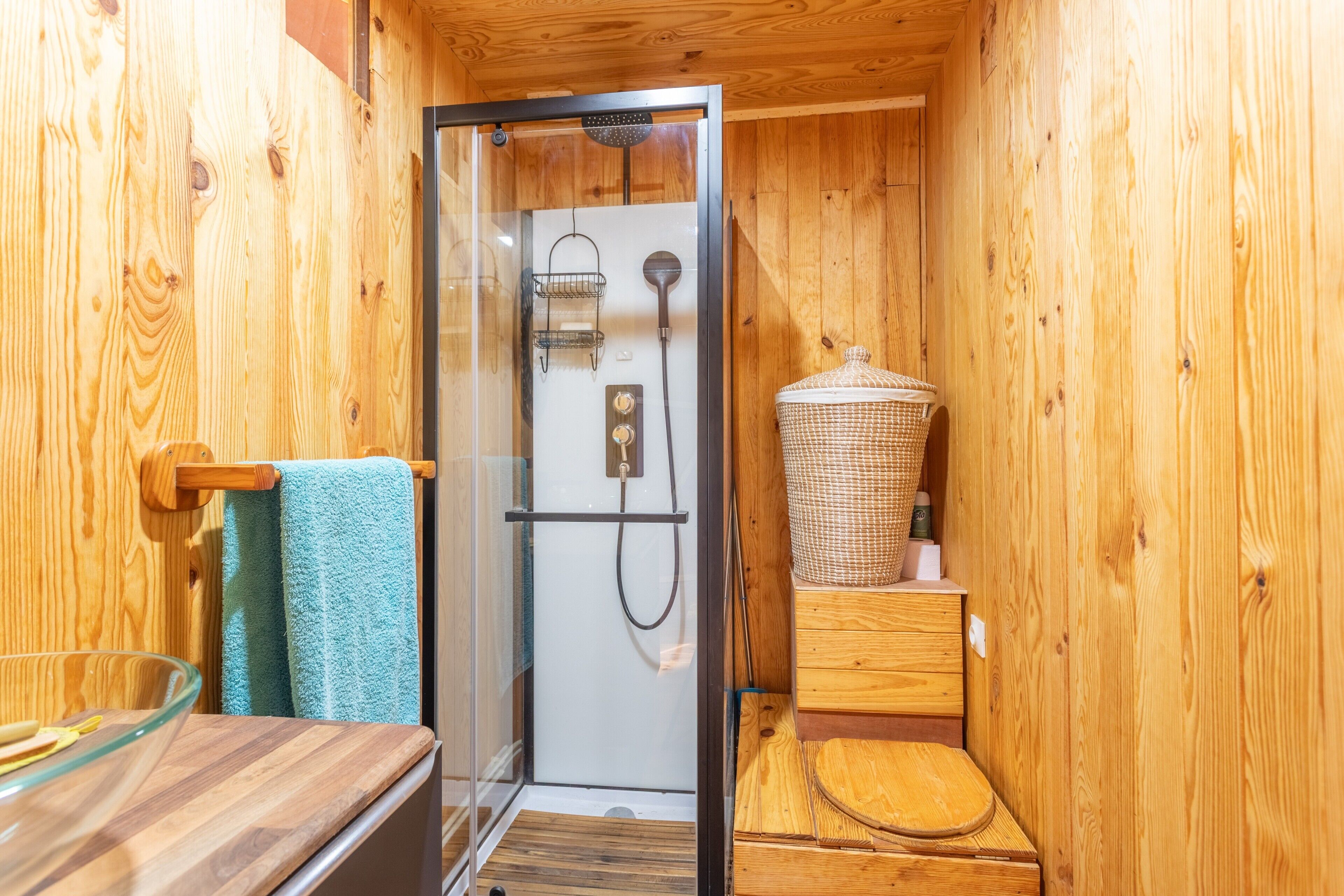 Ein Badezimmer in einer Holzhütte mit einer gläsernen Dusche, einer Holzbank und einem Korbwäschesammler.