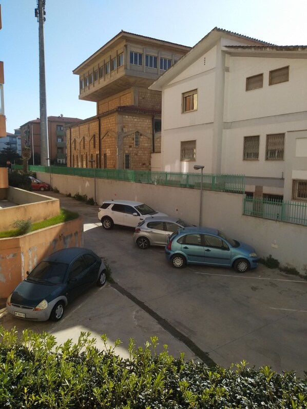 A parking area with several cars parked in front of buildings with a green fence and a clear sky. - Affittacamere Miri (Cagliari)