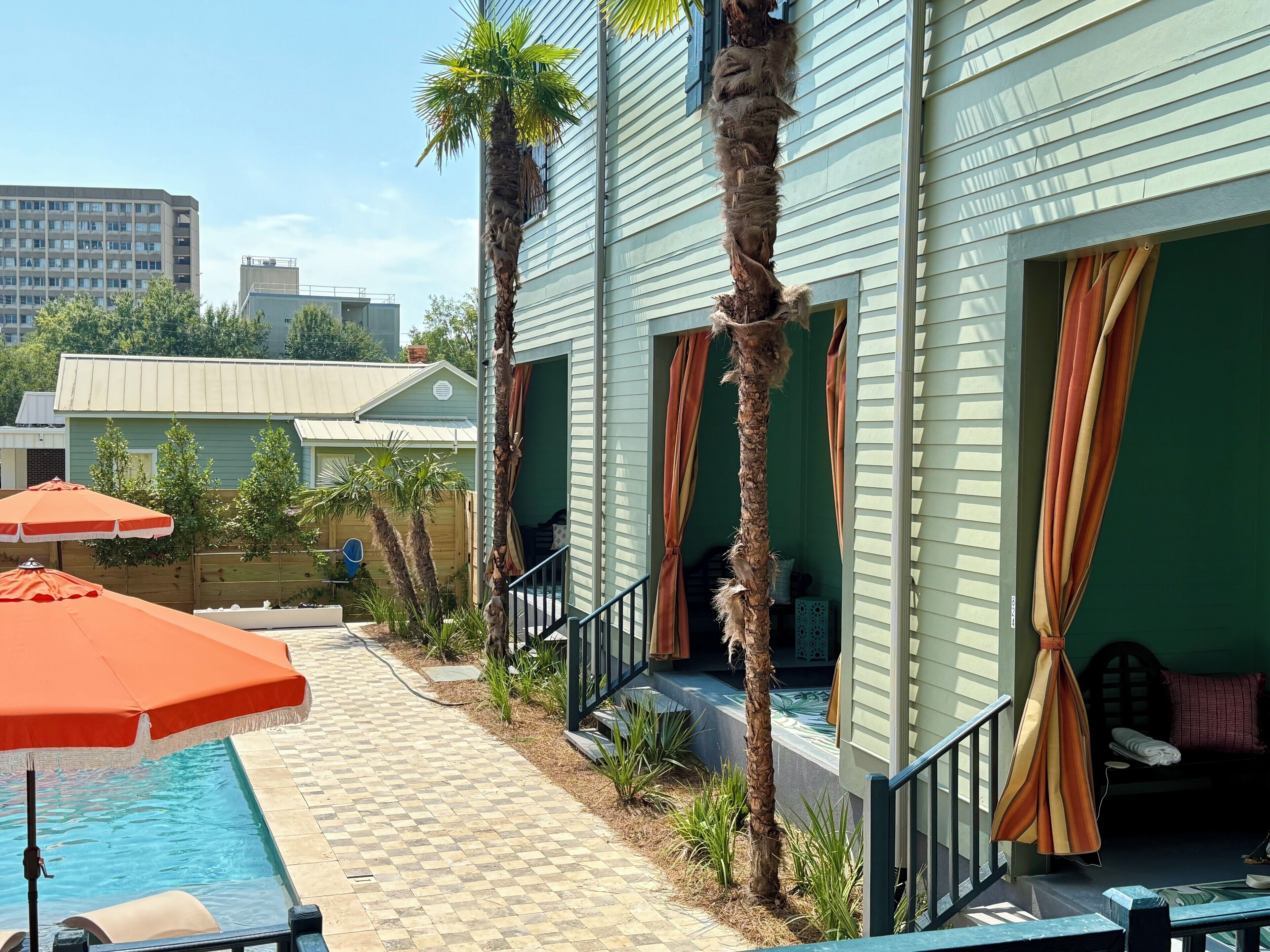 A pool area with a tiled walkway, palm trees, and a building with large windows and curtains.