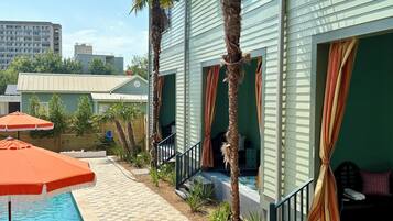 A pool area with a tiled walkway, palm trees, and a building with large windows and curtains.
