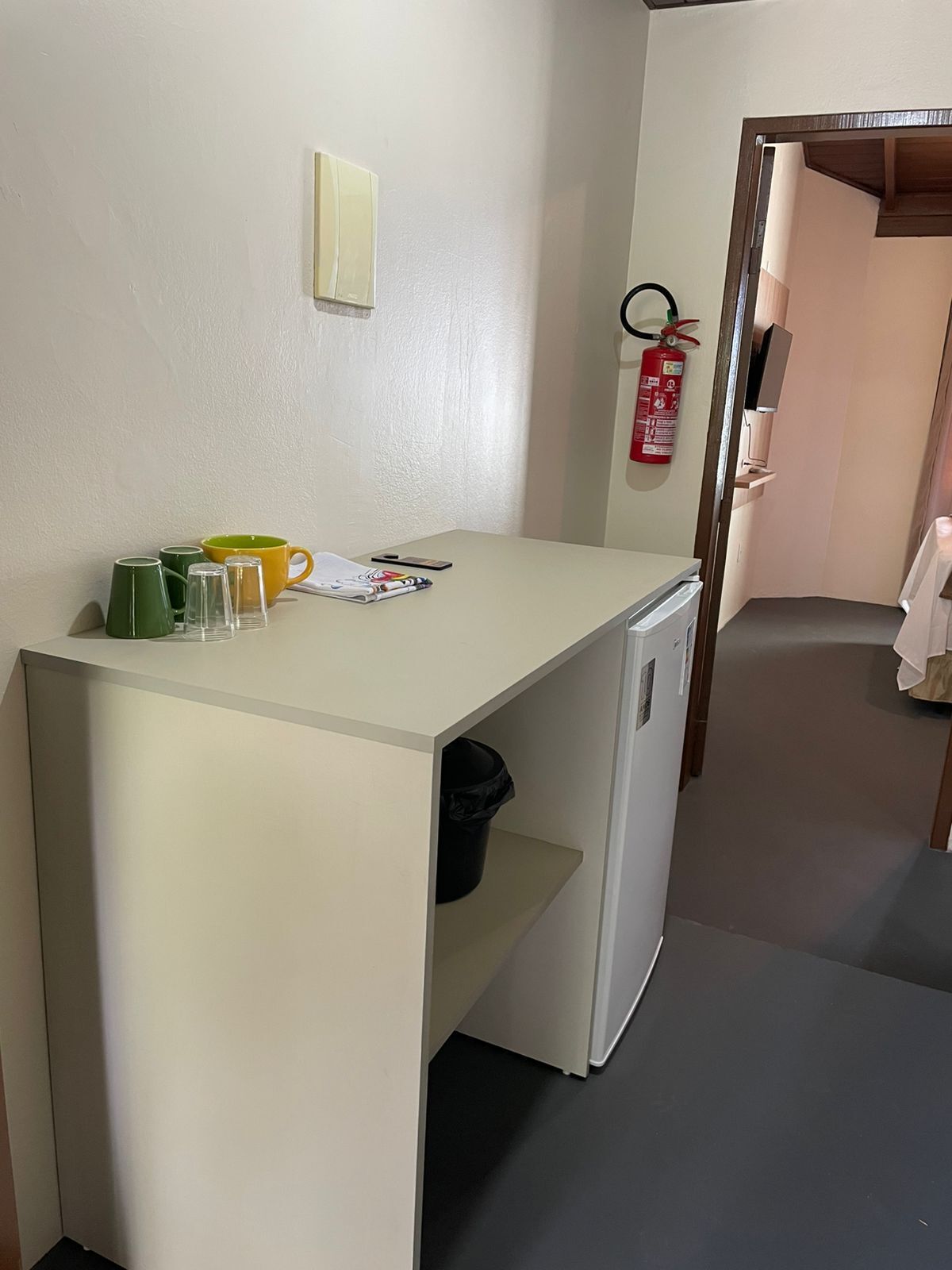 Een kleine keuken met een koelkast, een spoelbak en een aanrecht. Er staan groene en gele bekers, een tijdschrift en een brandblusser aan de muur.