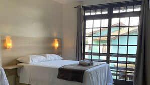 A hotel room with a bed, two bedside lamps, a window with a view of buildings, and a ceiling light. - Pousada Recanto Do Costão (Florianópolis)