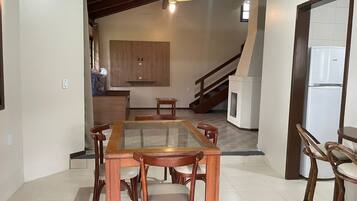 A dining area with a glass-top table and wooden chairs, a kitchen in the background, and a staircase leading to an upper floor.