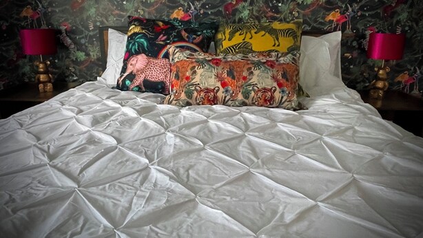Ein Bett mit weißer, gesteppter Bettdecke, farbenfrohen Kissen mit Tiermotiven und floralem Wandpapier.
