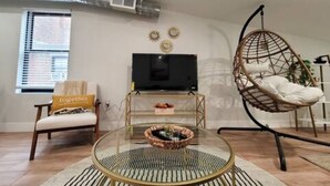 A living room with a television, a wicker chair, a glass coffee table, and a sofa with a 'together' cushion. - OTR by TQL+Studio+Queen Bed+50inTV+4th Fl+Sleeps 3ppl (Cincinnati)