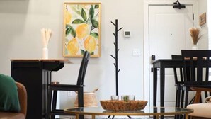 A living room with a glass coffee table, a painting of lemons, a coat rack, and a door. - OTR by TQL+Studio+Queen Bed+50inTV+4th Fl+Sleeps 3ppl (Cincinnati)