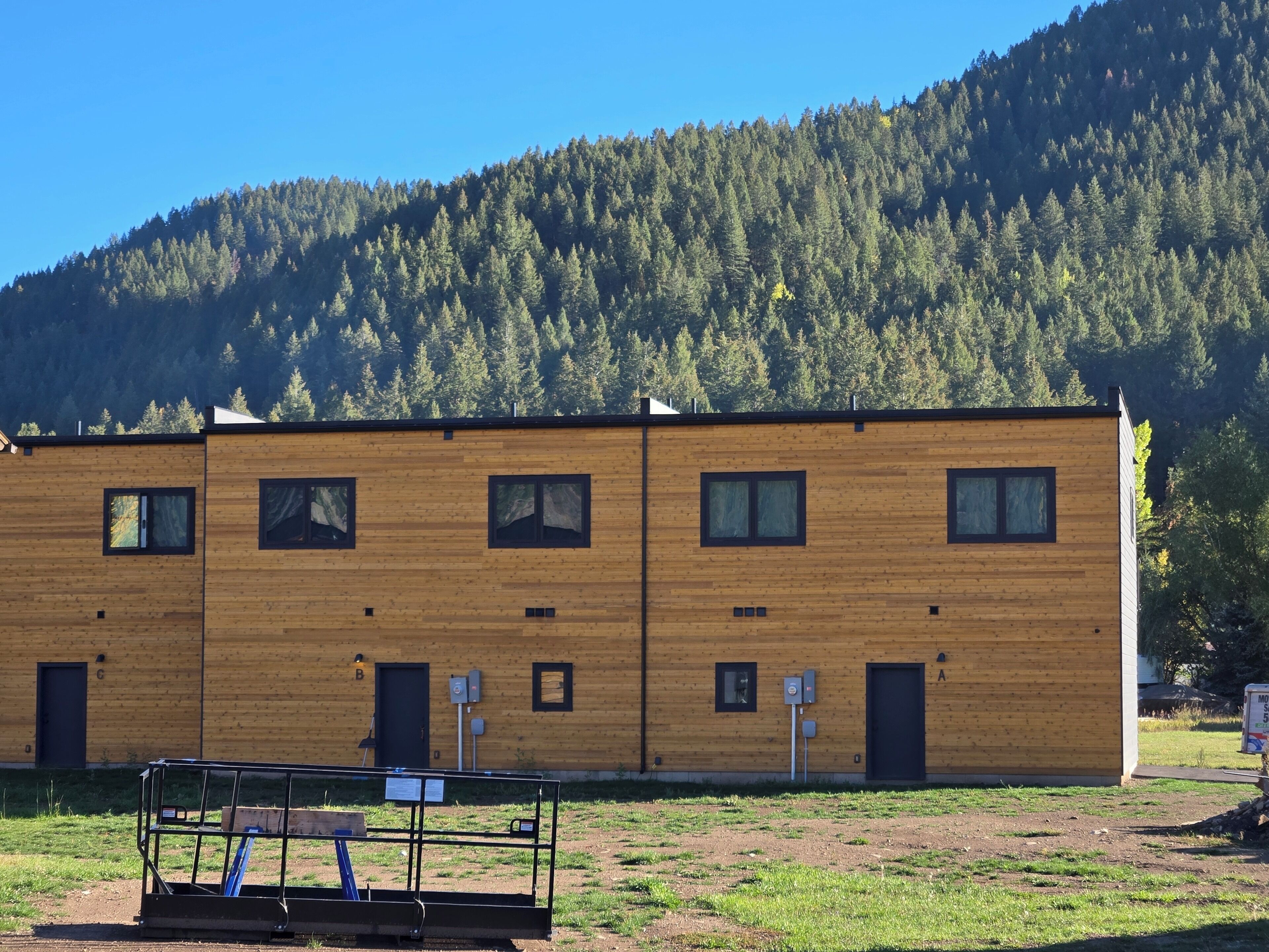 A two-story wooden building with multiple windows and doors, situated in a grassy area with a forested hill in the background.