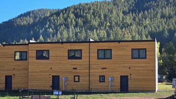 A two-story wooden building with multiple windows and doors, situated in a grassy area with a forested hill in the background.