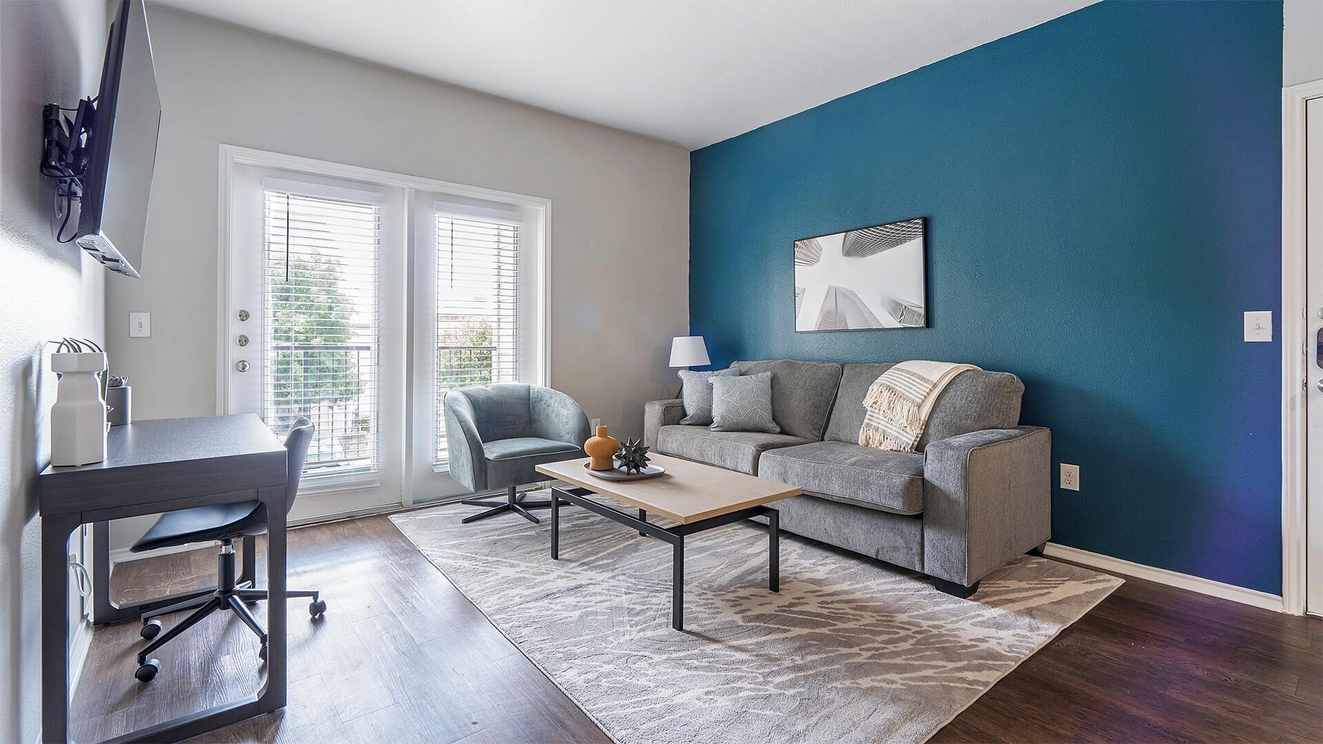 A modern living room with a grey sofa, a coffee table, and a desk with a chair. There are two windows with blinds, a blue accent wall, and a painting above the sofa.