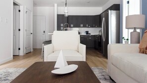 A modern living room with a white armchair, a coffee table with a decorative object, and a sofa. A kitchen is visible in the background with dark cabinets and a refrigerator.