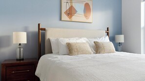 A neatly made bed with a white comforter and two decorative pillows, flanked by two bedside tables with lamps, against a light blue wall with a geometric painting above.