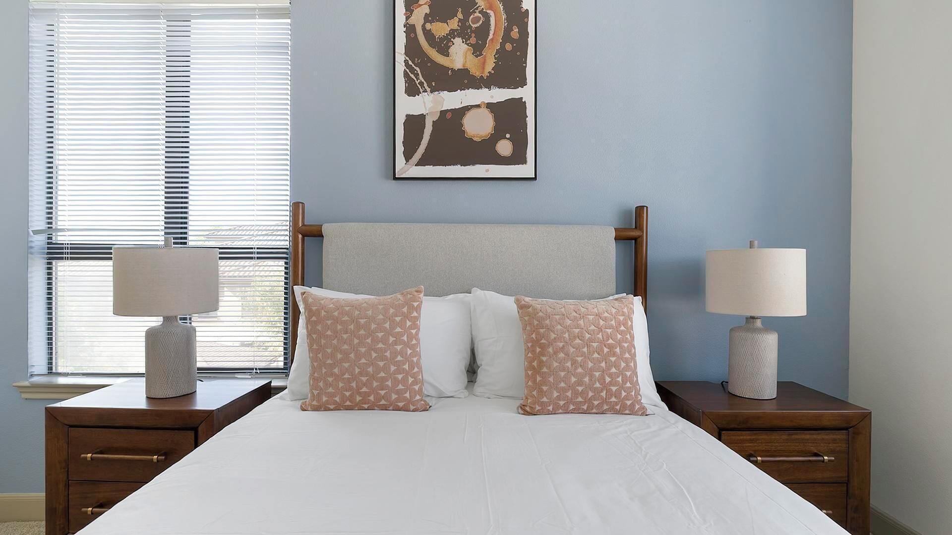 A bedroom with a bed, two nightstands, two lamps, a window with blinds, and a wall with a framed artwork.
