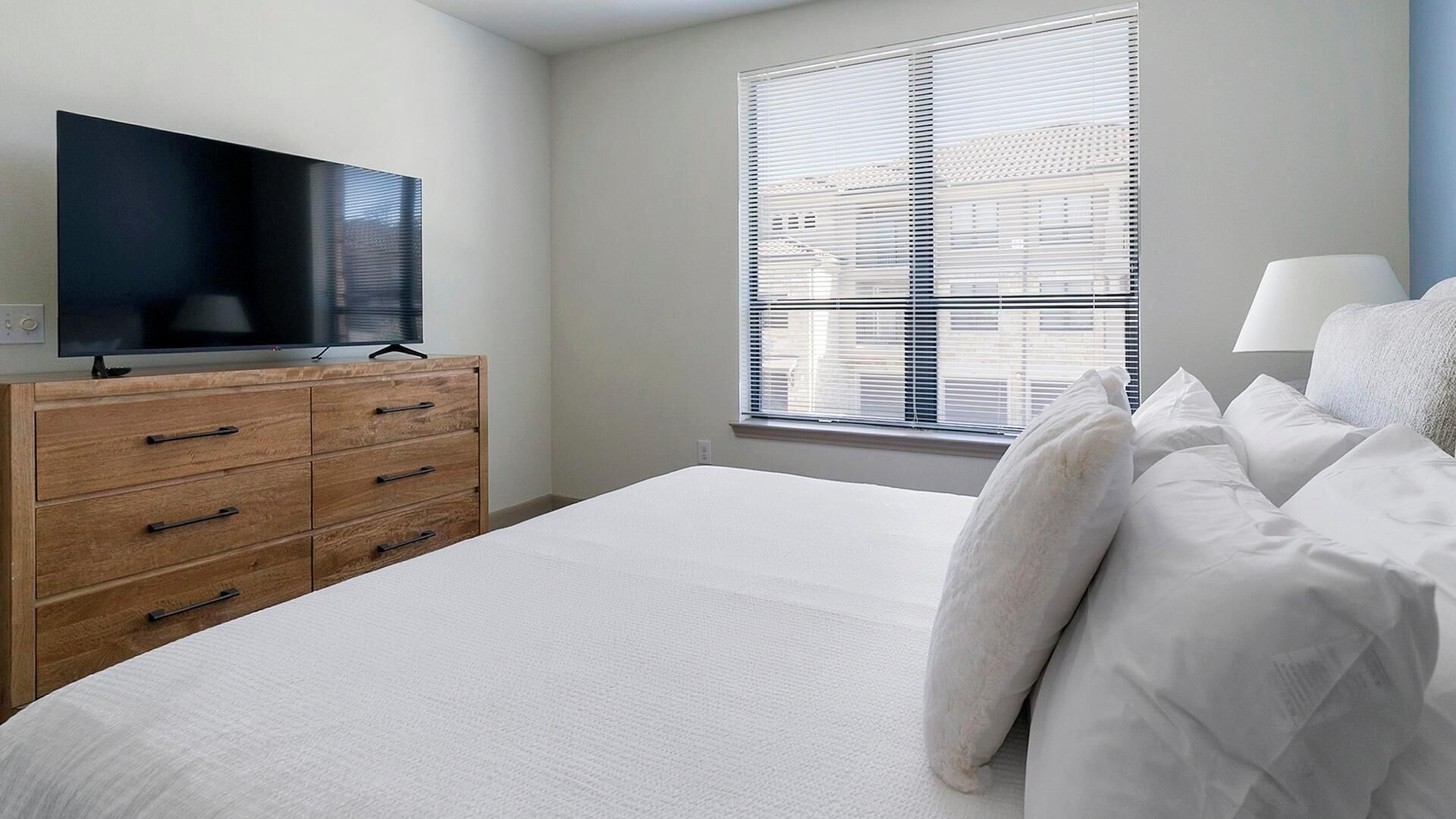 A bedroom with a bed, a wooden dresser, a television on top, and a window with blinds.