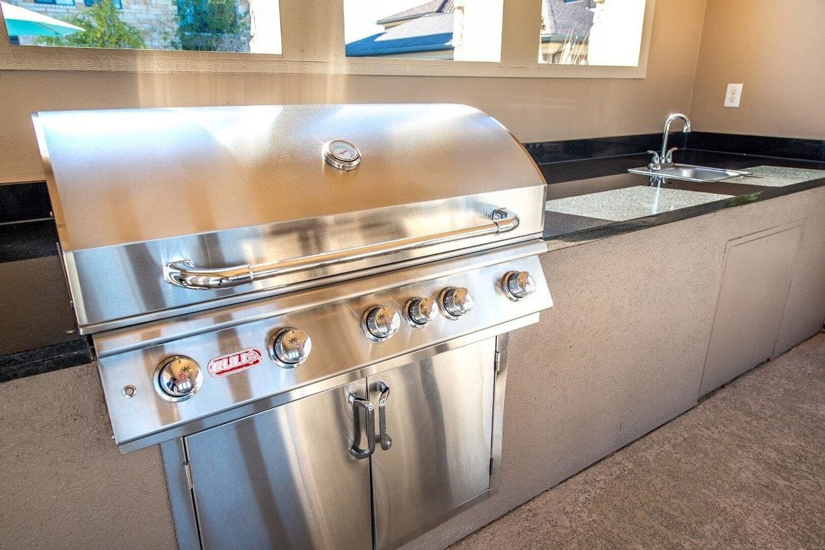 A stainless steel outdoor kitchen with a grill, a sink, and a window overlooking a residential area.
