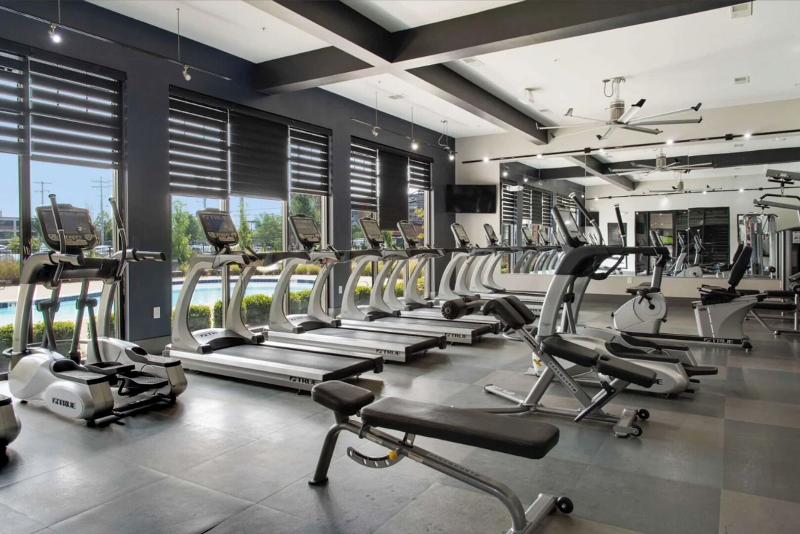 A modern gym with treadmills, ellipticals, and weight machines. Large windows offer views of a swimming pool and greenery.