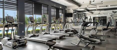 A modern gym with treadmills, ellipticals, and weight machines. Large windows offer views of a swimming pool and greenery.