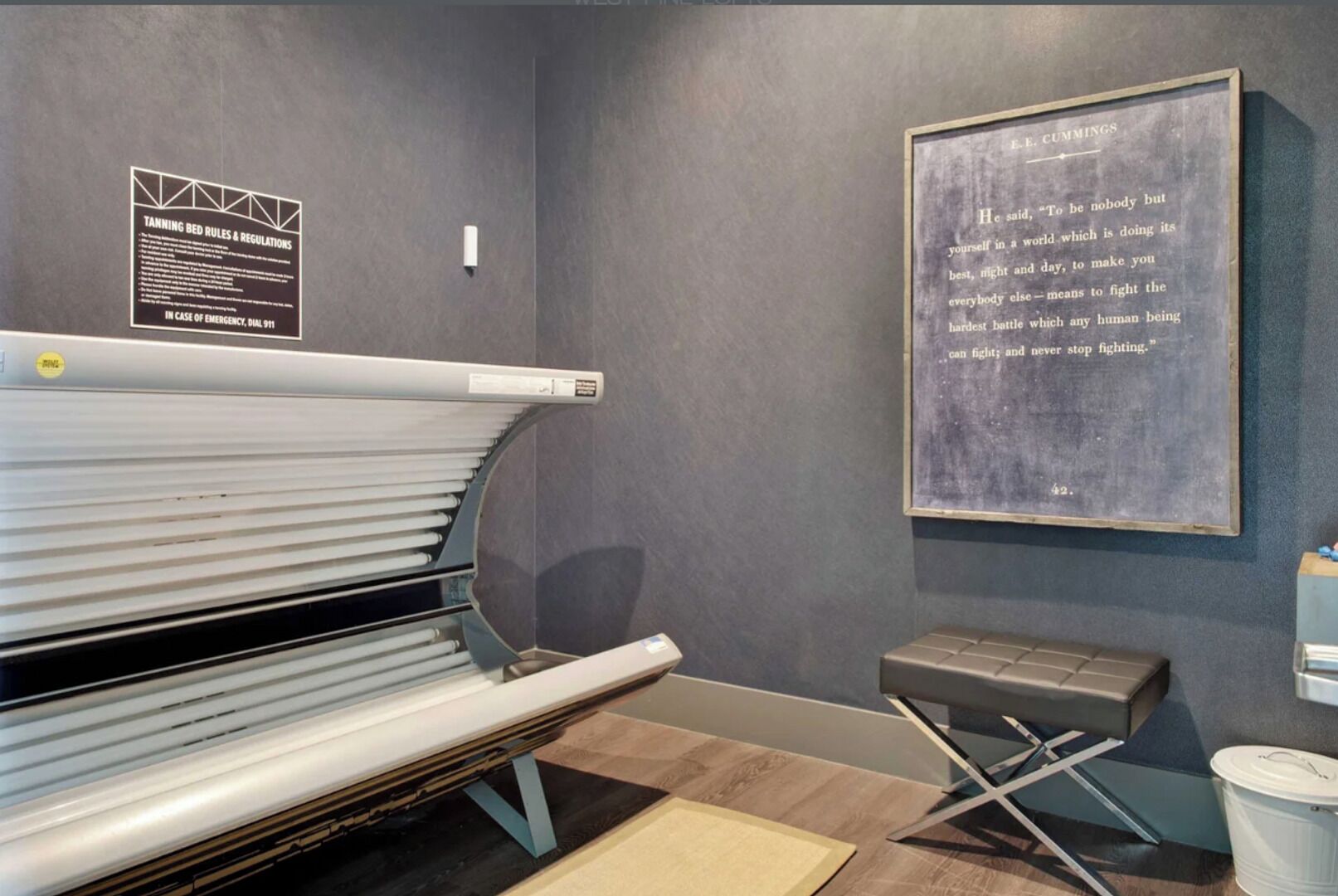 A tanning bed area with a sign detailing bed rules and regulations, a framed quote on the wall, and a small stool.