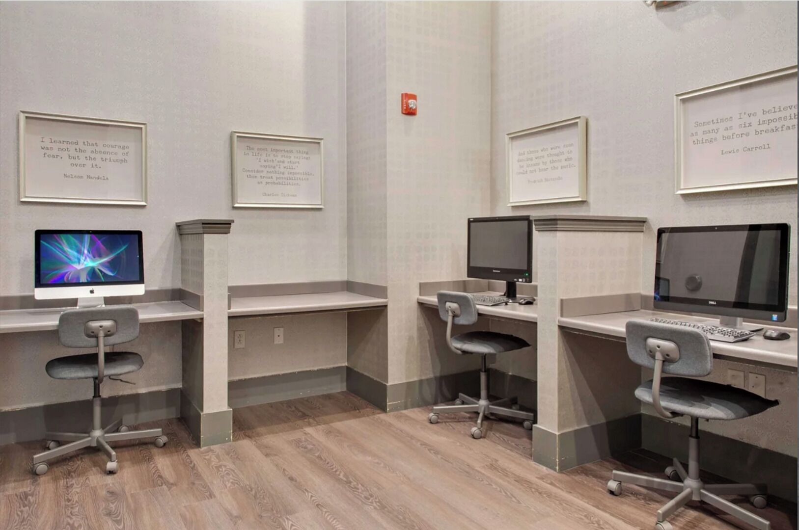 A modern office space with three desks, each with a computer monitor, keyboard, and chair. Inspirational quotes are displayed on the walls.