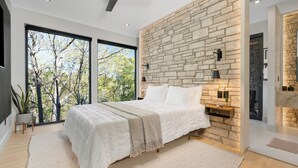 A modern bedroom with a large bed, stone wall, and large windows offering a view of trees. - The Lodge at Marble Creek. Modern Cabin Retreat /Hot tub/ Sauna/ Creek Access (Nicholasville)