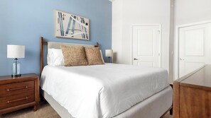 Una cama bien hecha con sábanas blancas, dos almohadas y cabecero, frente a una pared azul. Hay una obra enmarcada sobre la cama y una puerta blanca a la derecha.