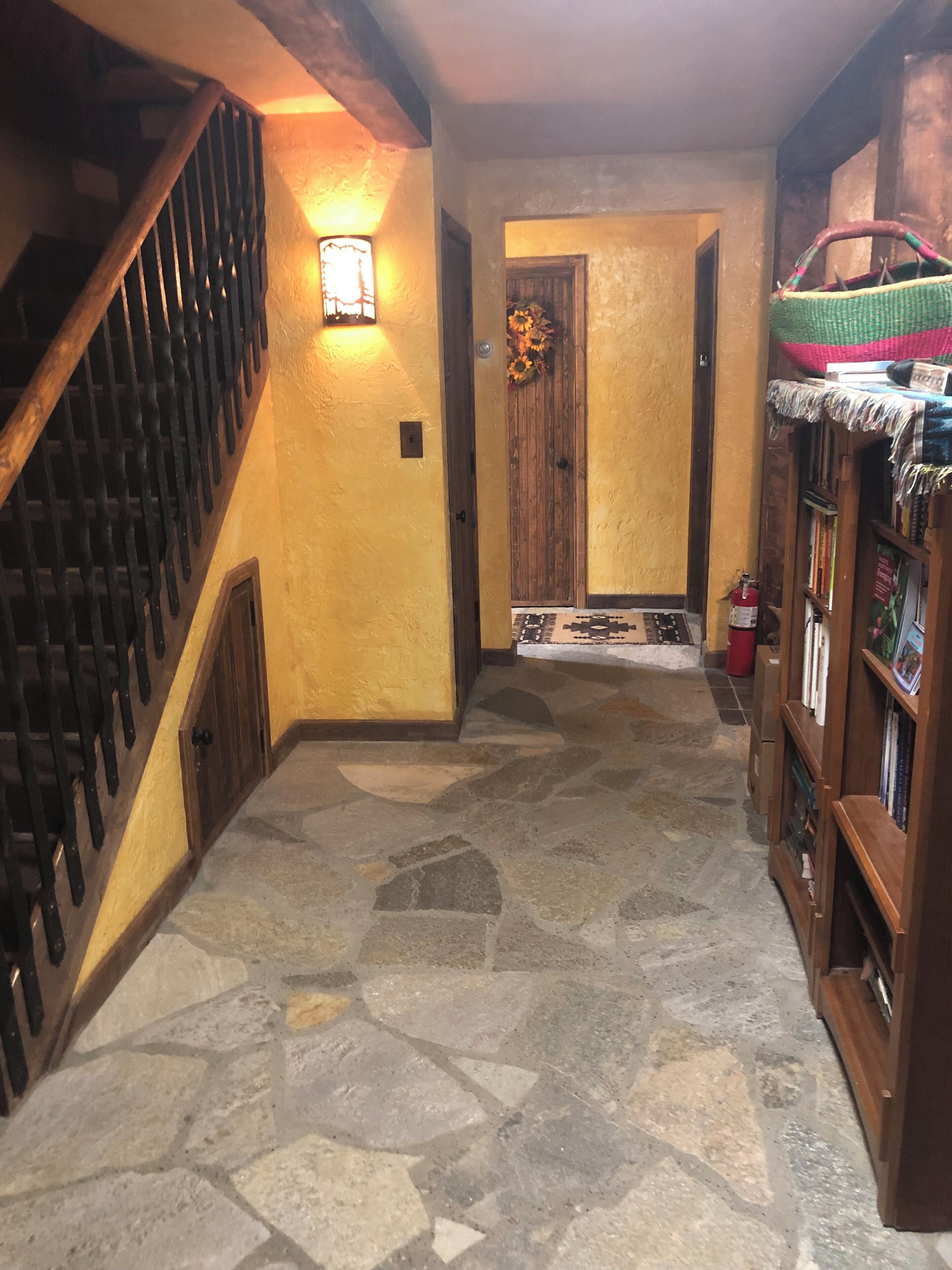 A stone-floored hallway with a wooden staircase on the left, a door with a floral wreath, and a bookshelf with various items on the right.