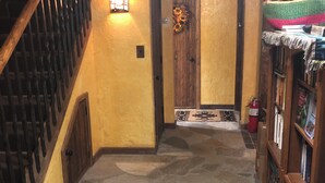 Un couloir au sol dallé de pierre, avec un escalier en bois sur la gauche, une porte ornée d’une guirlande de fleurs et une bibliothèque contenant divers objets sur la droite.