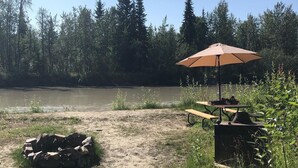 Un espace pique-nique équipé d’une table et d’un banc, d’un foyer et, en arrière-plan, d’une rivière.