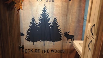 Un baño con cortina de ducha temática de bosque, que incluye siluetas de árboles y un alce, y el texto "WELCOME TO OUR NECK OF THE WOODS".