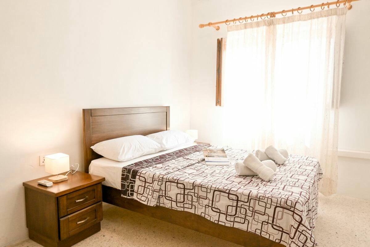 Een net opgemaakt bed met een bedsprei met een patroon, een nachtkastje met een lamp en een raam met dunne gordijnen.