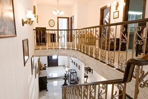 Una gran escalera con barandillas de metal ornamentadas, un candelabro y un suelo de mármol que conduce a una zona en el segundo piso con un conjunto de asientos.
