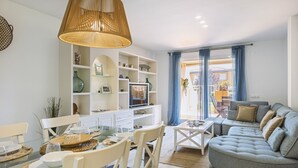 Ett modernt vardagsrum med ett glasbord, vita stolar, en grå soffa och ett stort fönster med gardiner.