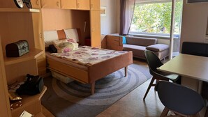 A compact bedroom with a bed, a desk, a chair, and a window with a view of greenery.