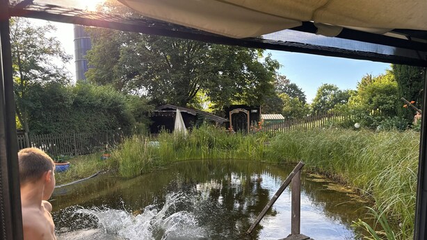 A wooden dock extending over a pond with water splashing, surrounded by greenery and a wooden fence.