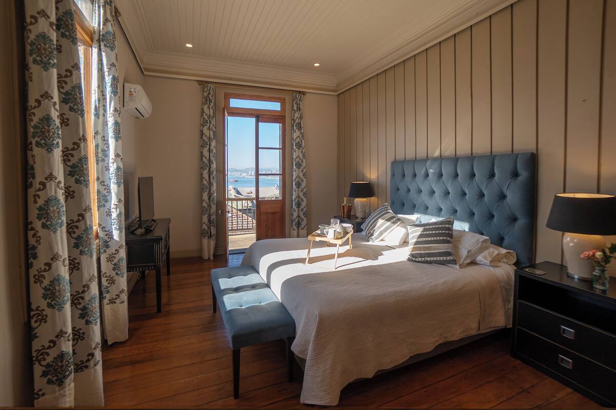 Une chambre à coucher avec un grand lit, deux tables de chevet, un banc, une commode et une vue sur l’extérieur.