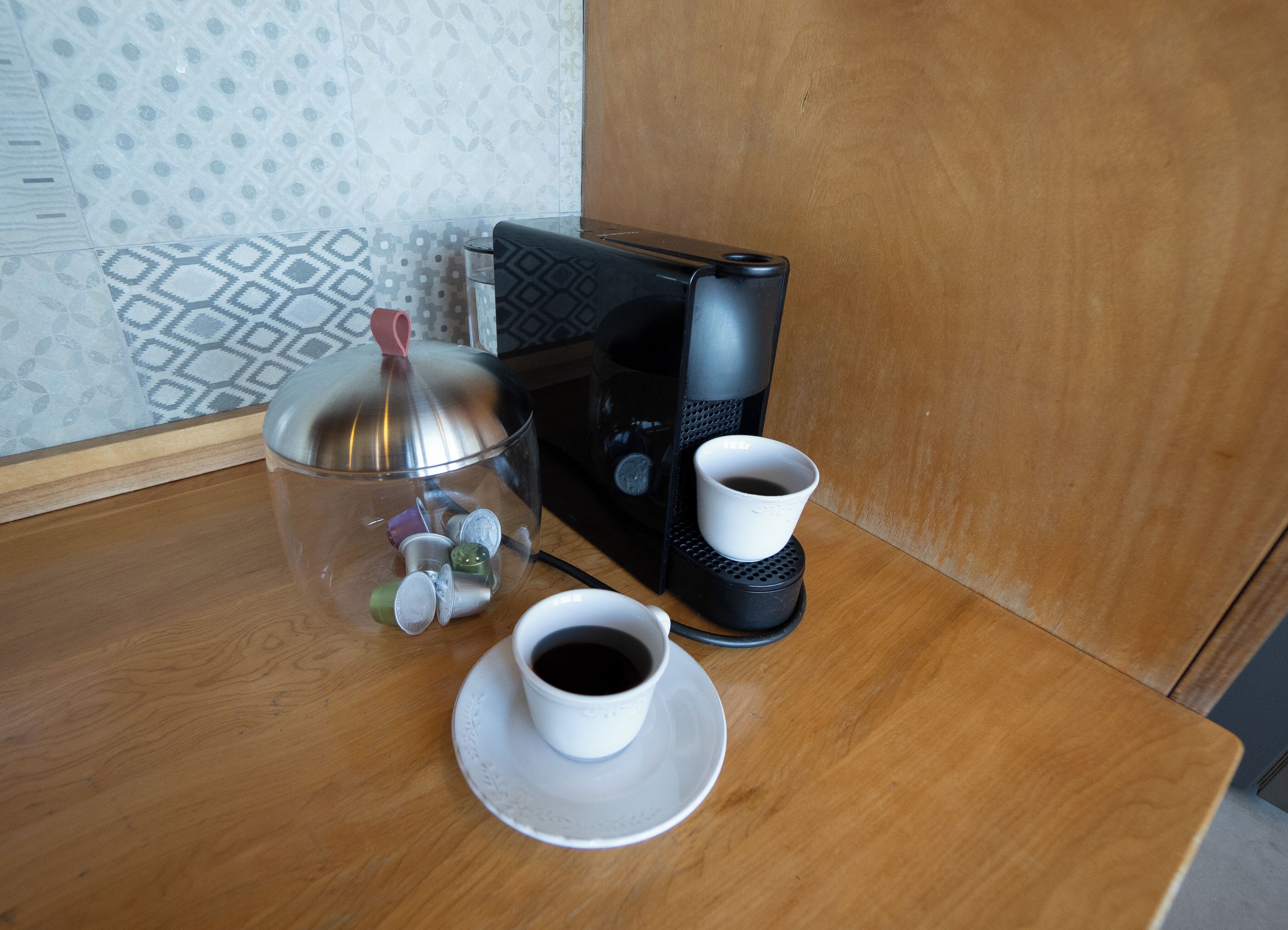 Una cafetera con una taza de café sobre una mesa de madera, un recipiente de vidrio con monedas y un adorno.