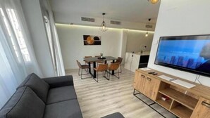 Un salon moderne avec un canapé foncé, un coin repas comprenant une table et des chaises, un téléviseur fixé au mur et un meuble en bois.