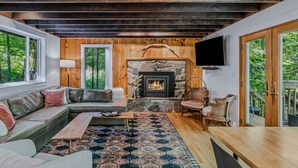 Ein gemütliches Wohnzimmer mit Kamin aus Stein, Holzbalken, einem großen Fenster und einem Fernseher.
