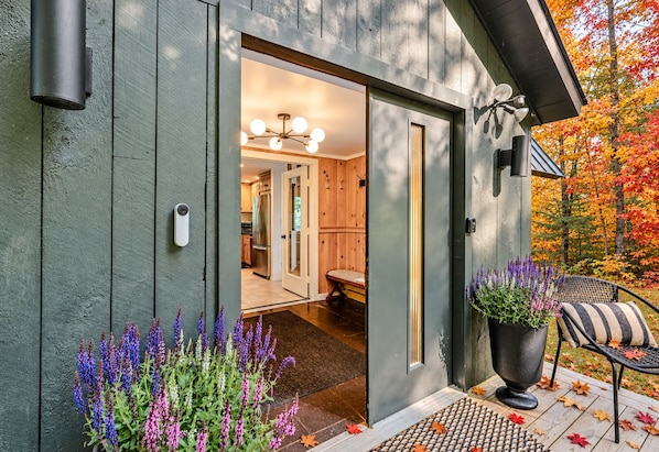 A wooden exterior with a door leading to a kitchen and living area, a porch with a chair and plants, and a view of autumn foliage.