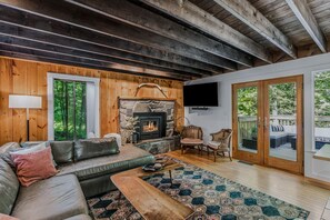 A cozy living room with a stone fireplace, wooden beams, a large window, and a television.