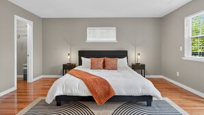A bedroom with a large bed, two bedside tables, a window with blinds, and a door leading to another room.