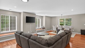 A modern living room with a gray sectional sofa, a flat-screen TV mounted on the wall, and a dining area with white chairs and a table.