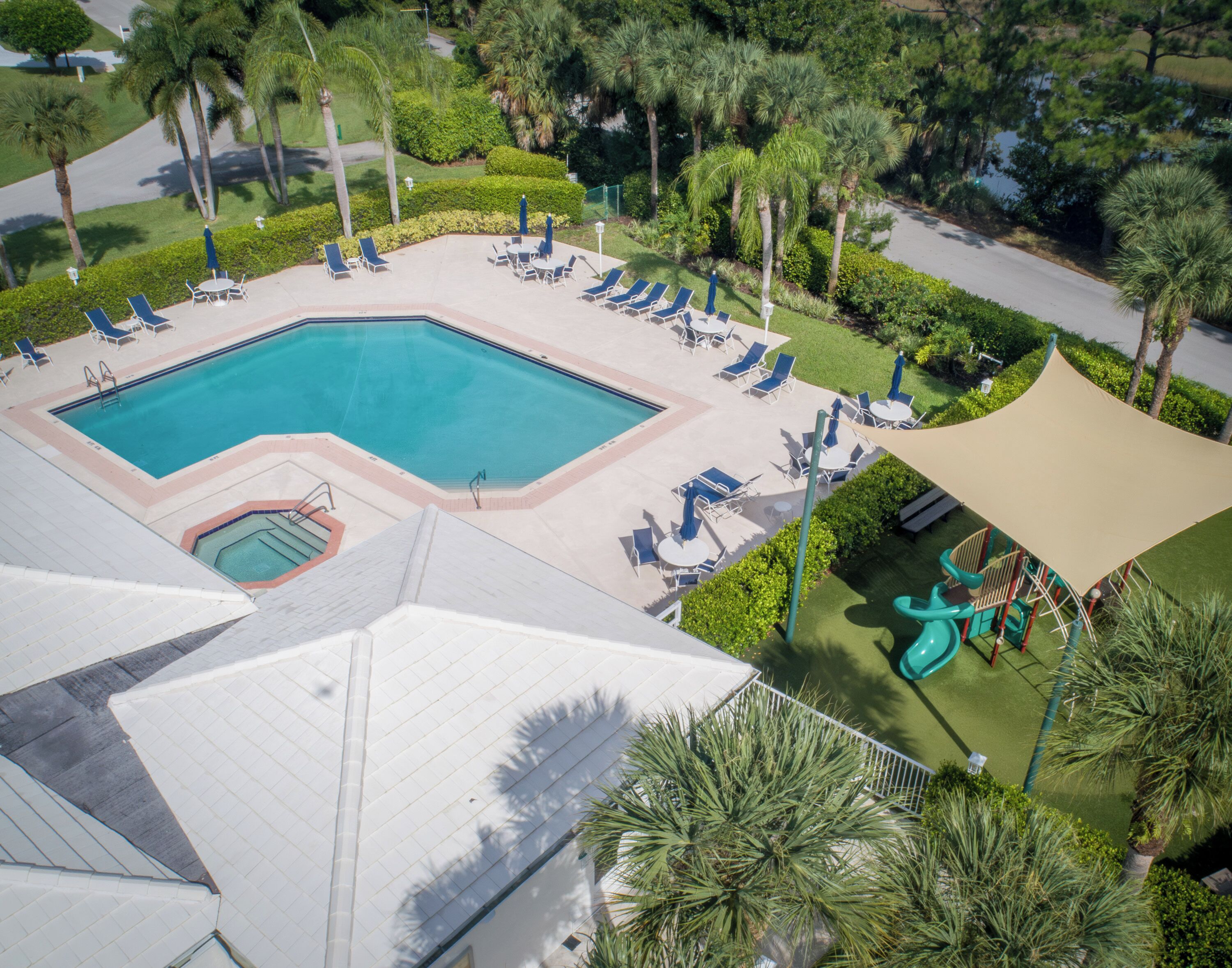 Vista aérea de um resort com uma grande piscina rodeada por espreguiçadeiras e guarda-sóis, área de recreação infantil e paisagismo bem cuidado.
