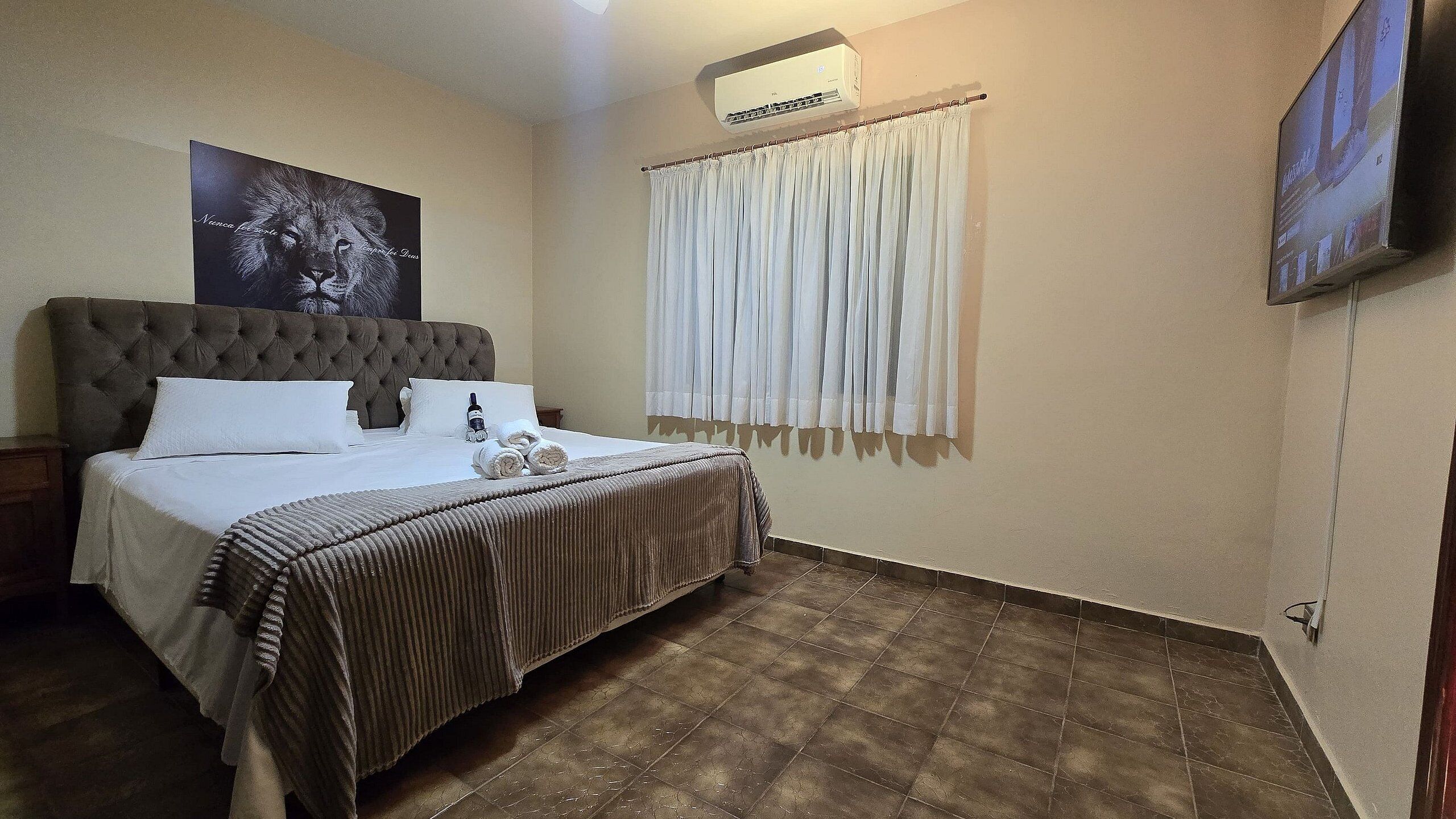 A bedroom with a tufted headboard bed, white linens, a striped bedspread, a mounted air conditioner, a large window with white curtains, and a flat-screen TV on the wall.