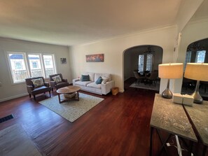 A spacious living room with hardwood flooring, a white sofa, two armchairs, a coffee table, and a dining area with a chandelier.