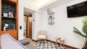 A modern hotel lobby with a grey sofa, a wicker chair, a small table with a remote, a wall-mounted TV, and a decorative plant. - Palm White Apartment, Vilamoura, Algarve (Quarteira)