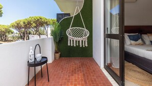 A balcony with a hanging chair, a small table with decorative items, a potted plant, and a view of the greenery and trees in the background. - Palm White Apartment, Vilamoura, Algarve (Quarteira)