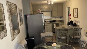 Un coin repas avec une table en verre entourée de chaises en métal, un réfrigérateur et des meubles de cuisine en arrière-plan.