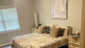 Une chambre avec un grand lit, une table de chevet, une commode, un ventilateur de plafond, une fenêtre avec des stores et un tableau au mur.