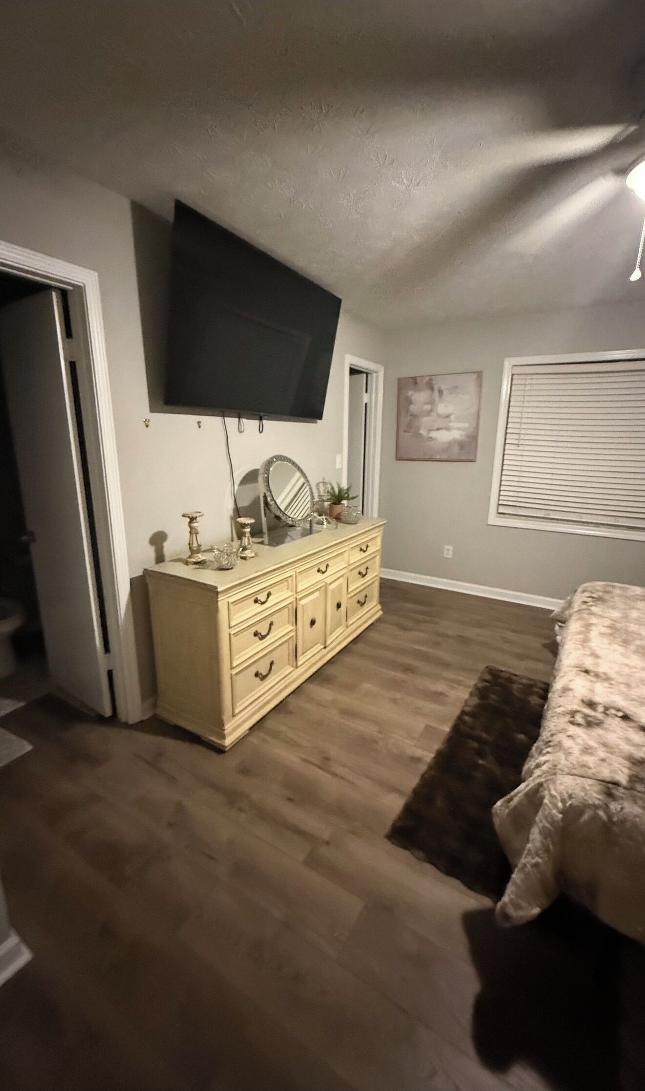 A bedroom with a large flat-screen TV mounted on the wall, a dresser with drawers, a bed with a patterned bedspread, and a window with blinds.