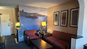 A hotel room with a sofa, armchair, and a table. There is a painting of a mountain landscape on the wall.